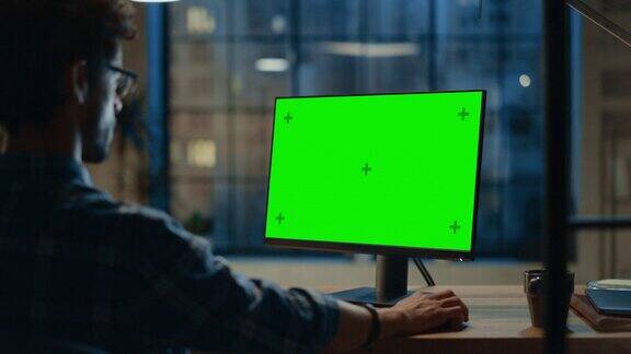 过肩:有创意的年轻人坐在他的办公桌上使用桌面电脑与模型的绿色屏幕晚上在时尚的办公室工作室与城市窗户景观