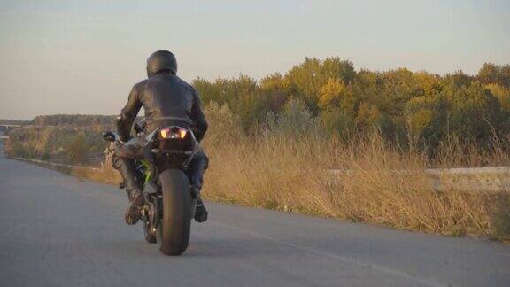 在乡村柏油路上骑着摩托车的男摩托车手的后视图在温暖的秋天年轻的摩托车手骑着现代运动摩托车在高速公路上疾驰近距离