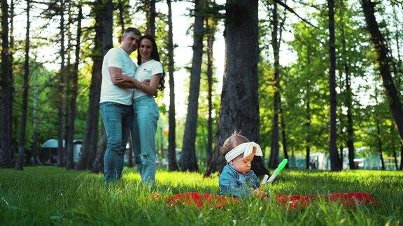 女婴坐在绿草地上的毯子上父母从远处看着她