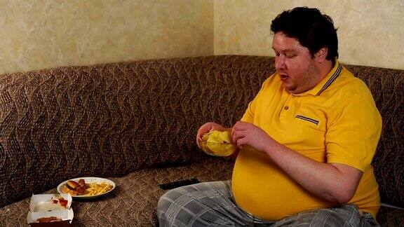 一个胖子坐在家里的沙发上看电视吃快餐