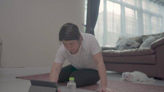 年轻的亚洲女人看着平板电脑在家里的客厅里和小狗一起锻炼