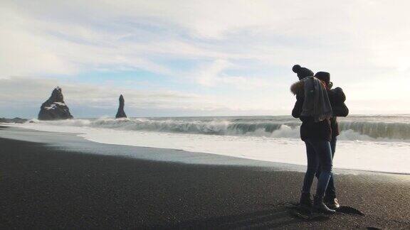 年轻的潮人夫妇在冰岛的火山黑沙滩上跑步玩得很开心慢镜头Reynisfjara海滩Reynisdrangar巨怪脚趾山Vik