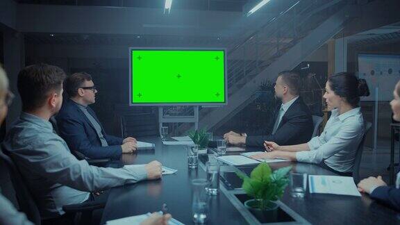 深夜在公司会议室:董事会高管和商人坐在谈判桌上谈论和使用绿色模拟屏幕墙电视视频电话会议
