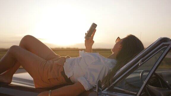 一名年轻女子躺在汽车引擎盖上使用智能手机