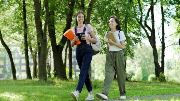 两个微笑的女大学生亚洲和高加索人背着背包课本和活页夹走过绿色的公园在阳光明媚的夏日交谈全程跟踪拍摄