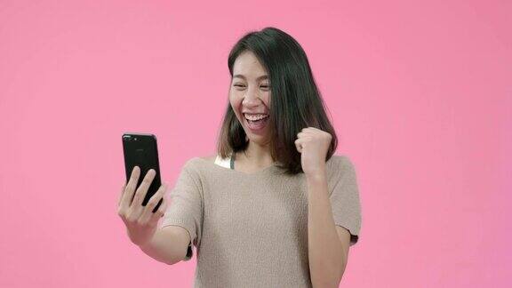 年轻的亚洲女子用智能手机查看社交媒体感觉快乐的微笑在休闲服装在粉色背景的工作室拍摄快乐的微笑可爱的快乐的女人庆祝成功