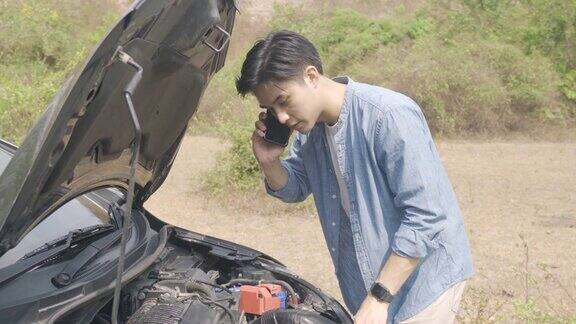 亚洲年轻的不快乐的男人在打开的引擎盖坏了的汽车前打电话在乡村路打电话在旅途中路上的破车