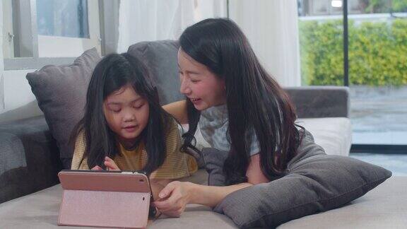 年轻的亚洲家庭和女儿喜欢在家里使用平板电脑日本妈妈和小女孩躺在家里客厅的沙发上看电影有趣的妈妈和可爱的孩子玩得很开心