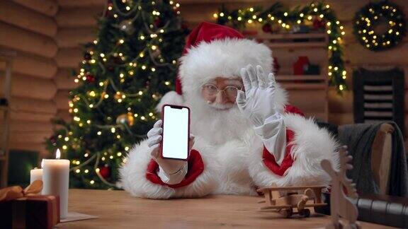 在装饰华丽的室内圣诞老人展示了一个空屏幕的智能手机并微笑着向相机挥手