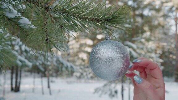 靠近女性手触摸银球挂在圣诞树外面白雪覆盖的森林里年轻女子的双手在新年树上挂着球玩具