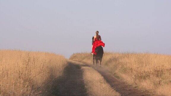 穿着红色长裙的年轻女孩骑着黑马在干燥的草原上