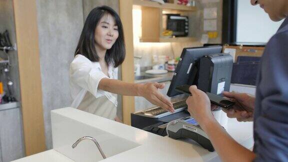 在咖啡馆里一位美丽的亚洲女人正在为一位通过非接触式手机向信用卡系统付款的顾客制作外卖咖啡