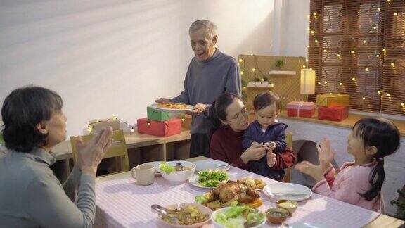 亚洲的祖父端上披萨并把它放在感恩节的餐桌上同时一起在公寓庆祝传统节日
