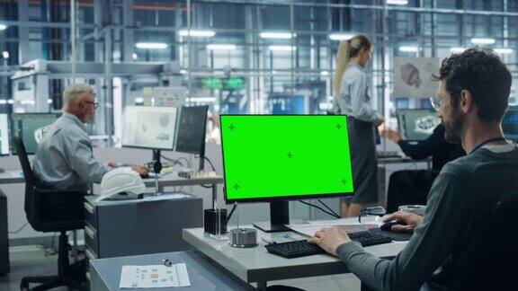 汽车工厂办公室:男汽车工程师坐在他的办公桌上工作的绿色屏幕色度键电脑自动化机械臂装配线制造中宽弧光照射