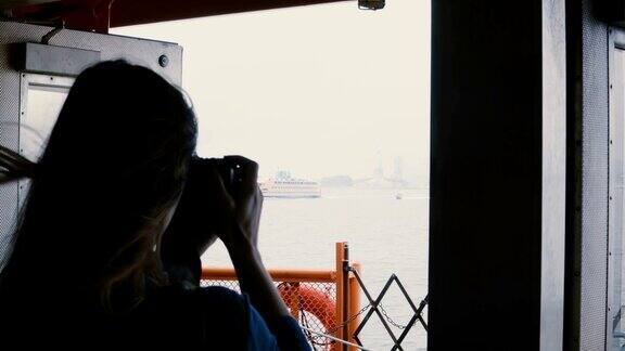 后视图年轻女游客在一个阴天的自由女神像的旅游船上用相机拍照
