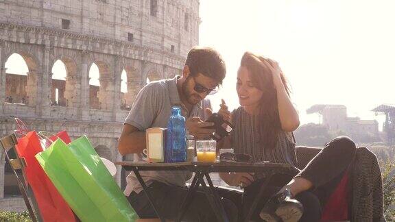 一对快乐的年轻游客夫妇在日落时分用智能手机自拍坐在罗马斗牛场前的酒吧餐厅里带着咖啡购物袋笑着玩