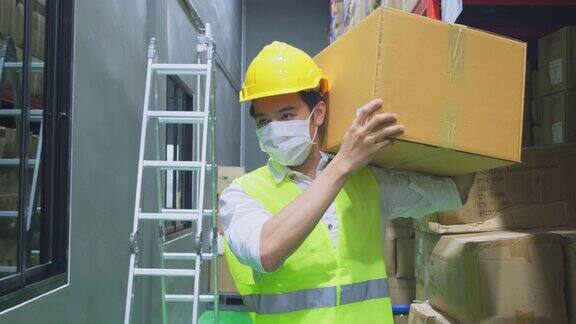 新冠肺炎流行期间亚洲男性工人戴口罩工作年轻人带着安全帽、背心搬运产品箱放到物流和出口工厂的货架上