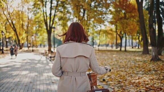 后视图不知名的红发女人推着她的巡洋舰自行车走在城市公园的瓷砖小路秋天的阳光灿烂的日子跟踪镜头慢动作