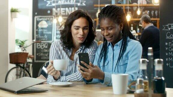 两个漂亮的女朋友坐在咖啡店聊天一个在她的智能手机上看照片他们都笑了在背景时尚的建立