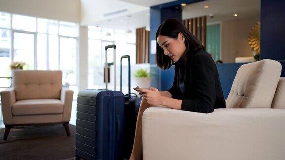 在一家豪华度假酒店的大厅里亚洲女商人穿着正装带着滚轮行李箱一边在智能手机上输入信息一边等着一位同事