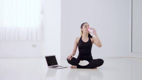年轻漂亮的亚洲女性在运动后喝水休息身穿黑色西装的女子在通过笔记本电脑观看在线教程完成瑜伽运动后坐在健身地板上喝着水