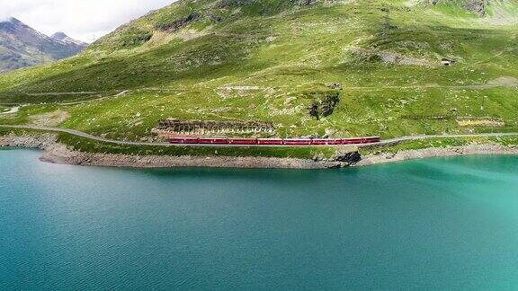 一列红色火车在瑞士山脚下的一个蓝色湖泊附近经过的无人机鸟瞰图美丽的风景下的蓝天4K