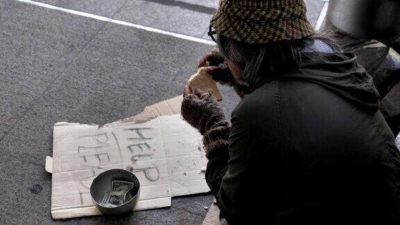 一个无家可归的老人穿着脏衣服一边吃着面包一边坐在街上寻求帮助