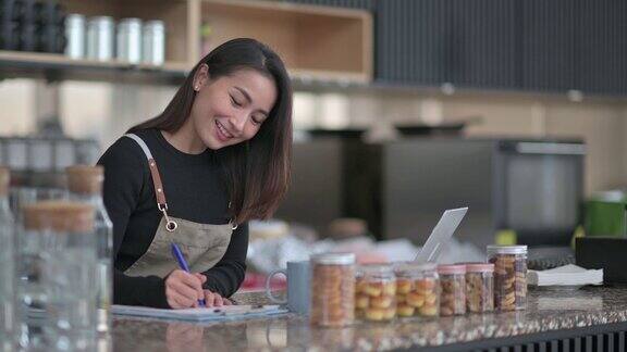 亚洲华人咖啡馆女老板与围裙使用笔记本电脑在咖啡馆柜台