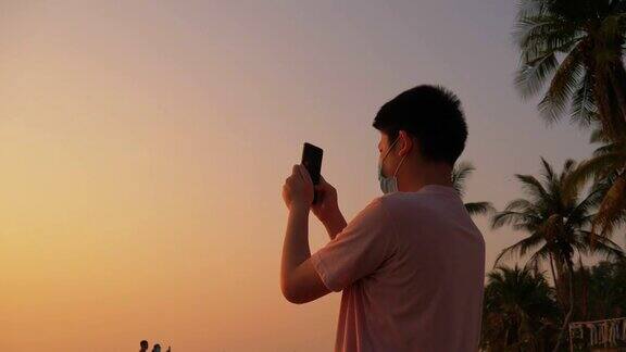快乐亚洲男孩的剪影拍照和自拍的智能手机在海滩上日出度假生活理念