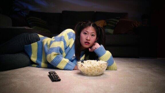 年轻漂亮的亚洲女人惊讶地躺在客厅地毯上吃着爆米花