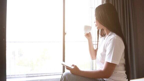 亚洲妇女坐在床上喝一杯咖啡同时使用手机在卧室在早上