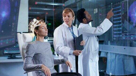 戴着脑电波扫描耳机的女人坐在椅子上两名科学家在监督和查看数据在现代大脑研究实验室监测显示脑电图阅读和大脑模型