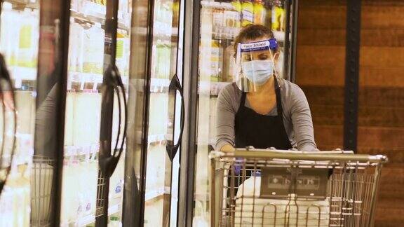 现实生活:超市女员工戴着防护面罩和口罩用一加仑瓶装的牛奶填满冷藏货架