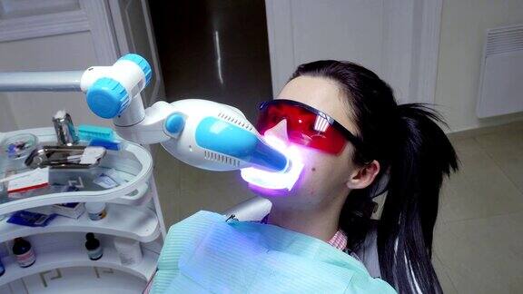 年轻的女人嘴上带着扩张器戴着红色防护眼镜正在进行紫外线美白一台在病人牙齿上运行的紫外线美白机