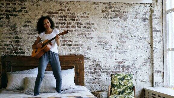 混合种族年轻有趣的女孩唱歌和弹吉他而有乐趣跳舞在床上在家