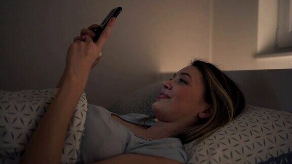 微笑的年轻女子躺在她的床上在她的手机在线约会应用聊天