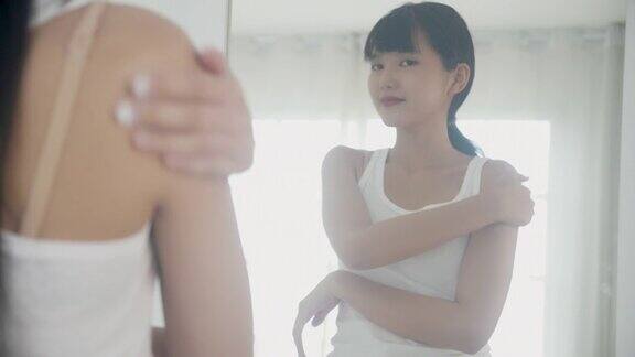 美丽的年轻亚洲女性在家里的浴室里看着镜子用化妆品霜或乳液润肤露和肩膀皮肤护理的卫生治疗和身体护理皮肤护理
