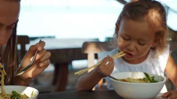 母女在露天的海滩咖啡馆用筷子吃面条汤