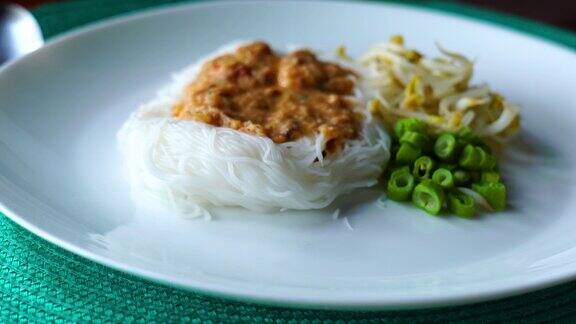 厨师将鱼辣椒酱倒在泰国米粉和蔬菜上放在蓝色的餐桌垫上的白色盘子里