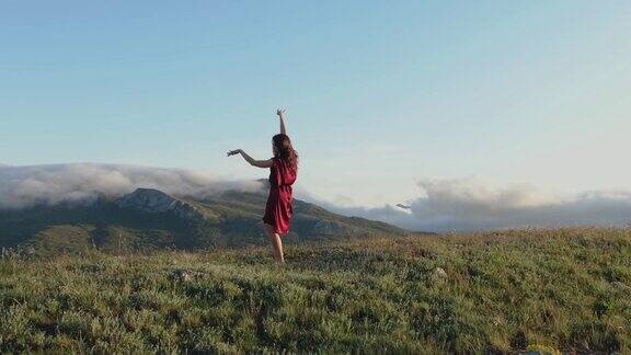 在白雾缭绕的山峦映衬下在夕阳的余晖中一位穿着红色连衣裙的年轻性感女子正在山顶上表演萨满舞蹈