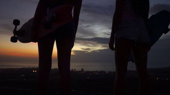 两个女孩站在路边手里拿着滑板看日落