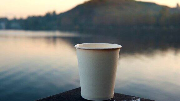 日落时分在水上的栏杆上放着一只装着热咖啡的白纸杯