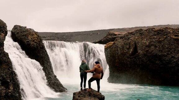 一对年轻夫妇在冰岛徒步旅行旅行的男人和女人站在强大的瀑布旁边举起手