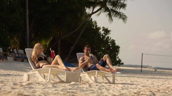 年轻的家庭夫妇日光浴和喝彩色鸡尾酒在热带海滩情侣在蜜月异国度假躺在海边的躺椅上的阳光度假