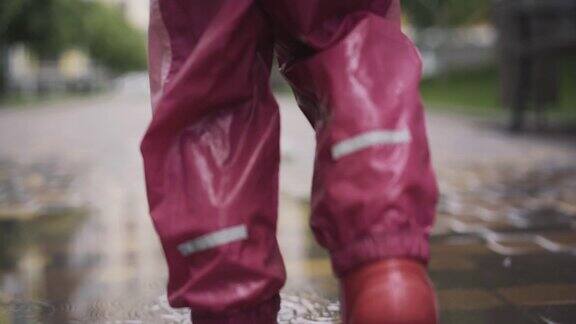 镜头跟随小女孩的双腿在水坑上行走在雨天沿着人行道散步的孩子在粉红色和红色衣服的背影童年休闲的生活方式