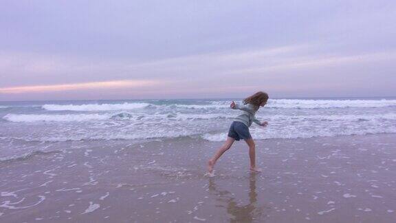 可爱的少女在日落时在海边跳舞和愚弄波浪