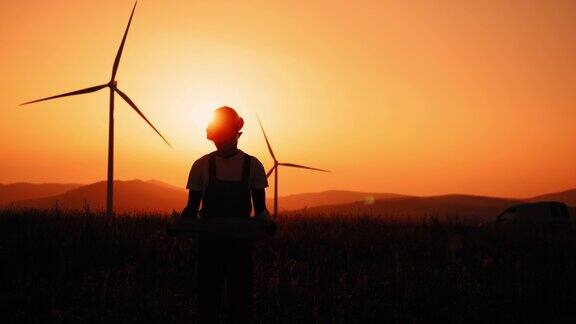 剪影男子工程师在安全帽站看着相机在晚上拿着生态站的大蓝图风力涡轮机一名非洲男子手里拿着蓝图站在装有风力涡轮机的农场里