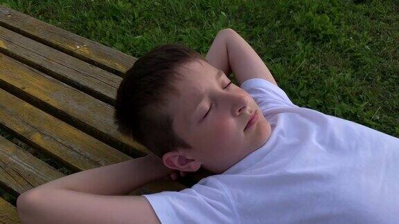 夏天的傍晚一个男孩闭着眼睛躺在长凳上在户外休息和做梦