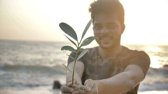 一个微笑的年轻人双手捧着一株绿色的小植物或样本对着冉冉升起的金色太阳