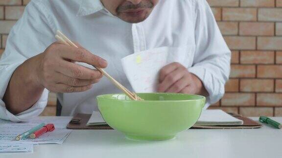 一名亚洲男子一边看书一边吃着绿色碗里的方便面